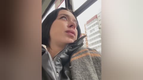 Media: A video captures a young woman with fair skin and dark hair, wearing a black leather jacket, looking contemplatively out a bus window. Modern buildings and overcast sky are visible.