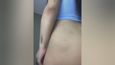 Media: Video of a woman's lower back and buttocks, wearing a light blue sports bra, showing stretch marks and a small tattoo on her right arm. The background is plain, beige walls.