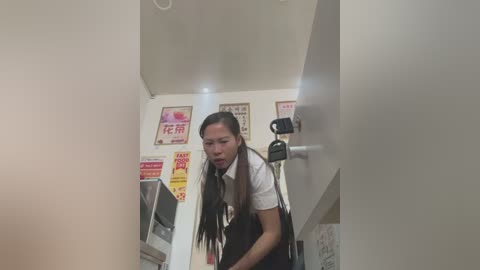 Media: A video of a young Asian woman with long black hair, wearing a white shirt and black skirt, looking distressed, in a dimly lit, cluttered room with posters and a door lock.