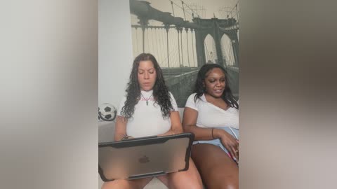 Media: Video of two Black women with medium skin tones, curly hair, and medium builds, wearing casual white tops and denim shorts, sitting on a couch, focusing on a laptop in front of them.