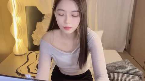 Media: A video of a young Asian woman with fair skin and long black hair, wearing a light gray off-shoulder top, sitting at a desk in a modern bedroom with beige walls, a white lamp, and a striped blanket.