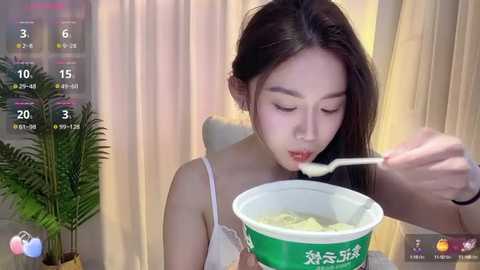 Media: Video of an East Asian woman with long black hair, wearing a white spaghetti strap top, eating noodles from a green bowl, set against a soft-focused background with a potted plant.