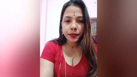 Media: Video of a South Asian woman with long, dark hair and fair skin, wearing a red blouse, a bindi on her forehead, and a gold necklace, indoors.