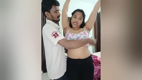 Media: Video of a South Asian couple in a bedroom; man in white polo shirt, woman in floral bra, stretching arms. Background includes bed with pink blanket.