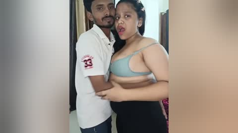 Media: A video of a South Asian couple standing close, smiling. The man, in a white polo, has medium skin tone, trimmed beard, and short hair. The woman, in a light blue bra and black pants, has medium skin tone and long black hair.