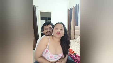 Media: Video of a man and woman in a bedroom; the woman, with long black hair and a floral top, leans forward, smiling, while the man, with a beard, is behind her, holding her waist.