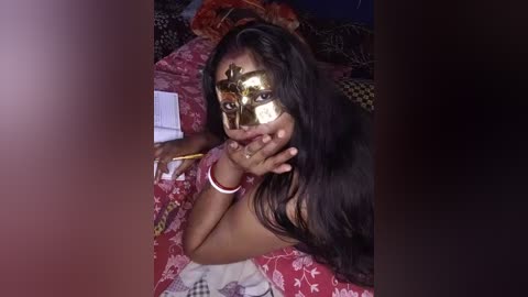 Media: Video of a young woman with medium brown skin, long dark hair, wearing a gold masquerade mask and red floral dress, lying on a red and white patterned bed, holding a book.