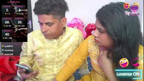 Media: A video of a young South Asian couple in traditional attire, the man in a yellow kurta, the woman in a yellow and gold sari, watching a smartphone together. Background shows a pink and white room.