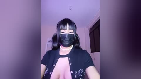 Media: Video of an East Asian woman with black hair and bangs, wearing a black face mask, black top, and pearl necklace, standing in a dimly lit room with white walls.