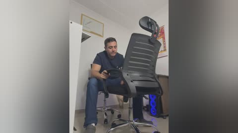 Media: Video of a man in a navy blue shirt, sitting on a black ergonomic office chair, typing on a laptop in a small, cluttered office with white walls and a framed certificate on the left.