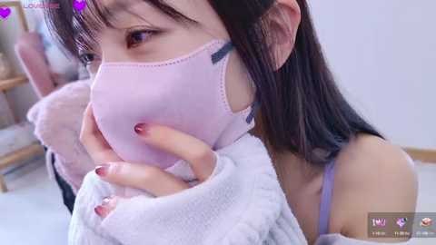 Media: A close-up video of an Asian woman with straight black hair, wearing a light pink mask and a light gray sweater, sitting on a bed, with a blurred background.