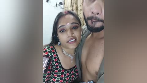 Media: Video of a young South Asian woman with a bindi, wearing a colorful top, posing with a shirtless man with a beard, in a domestic setting.