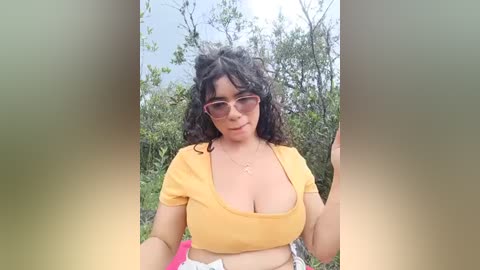 Media: Video of a light-skinned woman with curly black hair, wearing a yellow crop top, standing outdoors among green foliage.