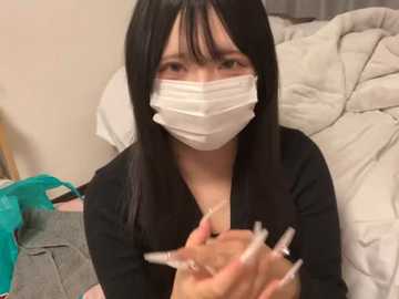 Media: Video of an East Asian woman with long black hair, wearing a white surgical mask, black top, and holding a syringe, sitting on a bed with white sheets and a teal garment in the background.