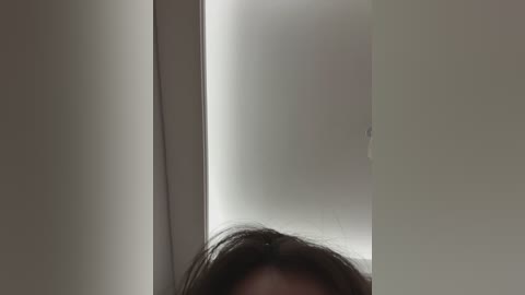 Media: Video of a person's face, partially obscured, with dark hair, viewed through a narrow, vertical gap in a white wall. The background is uniformly white, creating a stark, minimalist ambiance.