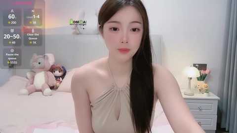 Media: A video of an East Asian woman with long brown hair, wearing a beige halter top, sitting on a bed with teddy bears, flowers, and a nightstand lamp in a softly lit room.