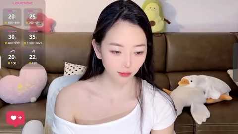 Media: A video of an Asian woman with long black hair and pale skin, wearing a white off-shoulder top, seated on a brown couch, surrounded by plush toys.