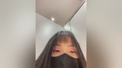 Media: Video of an Asian woman with straight black hair and bangs, wearing a black face mask, standing in a modern, minimalist room with white walls and recessed ceiling lights.