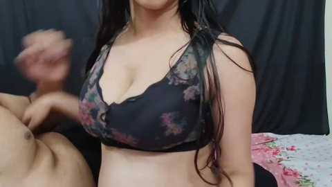 Media: Video of a young woman with long black hair wearing a floral-patterned bra, sitting on a bed with pink and white floral sheets, blurred man in background.