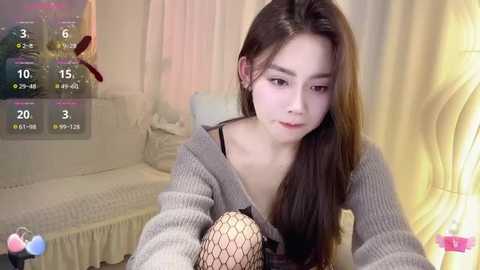 Media: A video of a young Asian woman with long brown hair, wearing a gray knitted cardigan, looking down at a baby doll, in a softly lit room with a bed and curtains.