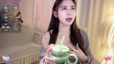 Media: Video of a young Asian woman with long black hair, wearing a gray sweater, holding a green straw cup, indoors with beige curtains and a couch in the background.