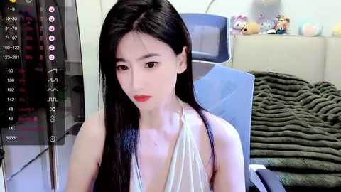 Media: A video of an East Asian woman with long black hair, wearing a sleeveless white dress, sitting indoors with a cluttered background featuring a blue chair, a stuffed toy, and a green blanket.