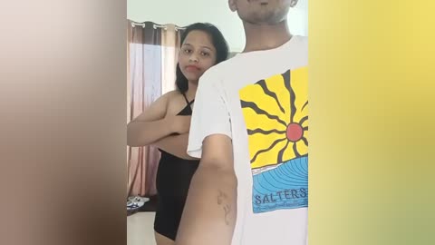 Media: Video of a young, light-skinned woman with medium-length black hair, wearing a black bra and shorts, standing close to a man wearing a yellow graphic t-shirt, in a brightly lit room with a window and curtains in the background.