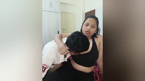 Media: Video of a young Asian woman with medium skin tone, medium build, and dark hair, wearing a black sports bra and black pants. She is hugging a man in a white shirt and red pants, both seated on a bed in a sparsely furnished room.