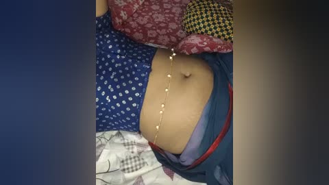 Media: Video of a partially clothed woman with light brown skin, wearing a blue patterned dress, a red belt, and a pearl necklace. She is lying on a bed with red, blue, and checkered patterned sheets.