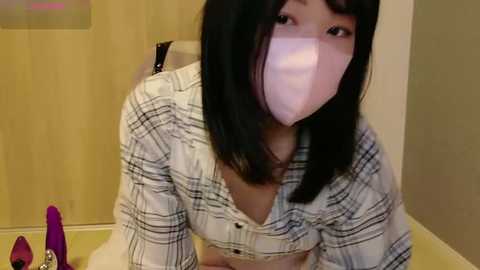 Media: Video of a young East Asian woman with straight black hair and a pale complexion, wearing a plaid shirt and a pink face mask, kneeling on a tiled floor, with a purple dildo and a small white bottle in the background.