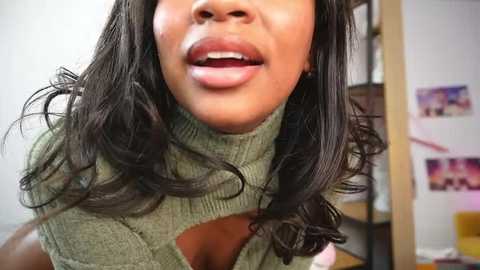 Media: Video of a smiling, light-skinned Black woman with long, wavy black hair, wearing a green turtleneck sweater, partially exposing her large breasts. Background shows a cluttered room with posters and a wooden shelf.