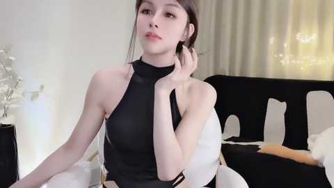 Media: Video of an Asian woman with light skin, dark hair, and delicate features, wearing a sleeveless black dress. She's seated on a white chair in a modern, dimly lit room with white curtains and a black and white cushion.