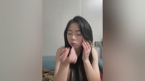 Media: A video of a young Asian woman with long black hair, wearing a sleeveless top, touching her face in a mirror reflection. The background features a light blue couch and beige wall.