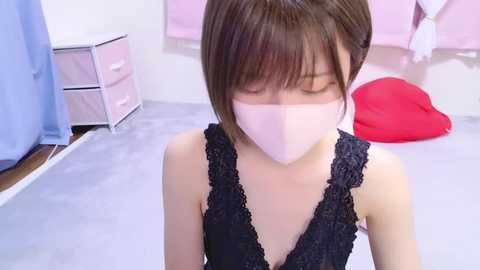 Media: A video of an East Asian woman with short brown hair, wearing a black lace dress and a pink face mask, kneeling in a pink and blue bedroom with drawers, a red cushion, and a pink wall.