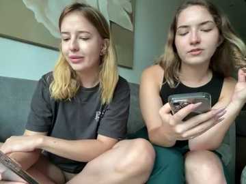 Media: Video of two young Caucasian women with light skin, sitting on a gray couch. One wears a gray t-shirt, the other a black sleeveless top. Both hold smartphones, appearing relaxed and engrossed.