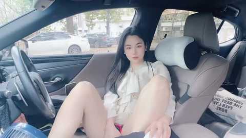 Media: Video of a young Asian woman with long black hair, wearing a white blouse and black shorts, sitting in a car with a parked white SUV visible outside.