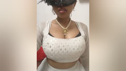 Media: Video of a woman with medium-dark skin, wearing a white sequined crop top that emphasizes her large breasts, black mask, and gold necklace. She's indoors, standing with a blurred background.