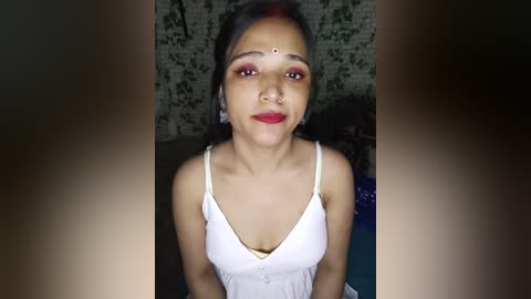 Media: A video of a young South Asian woman with medium skin tone, wearing a white spaghetti-strap top, red lipstick, and a bindi, sitting in a dimly lit room with a patterned brick wall.