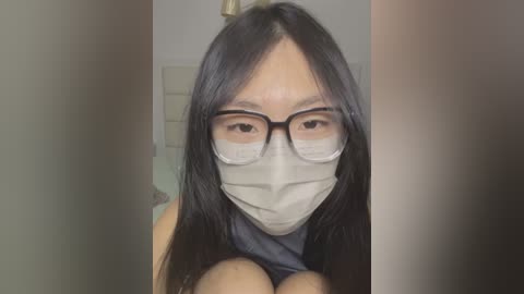 Media: Video of an East Asian woman with long black hair, wearing glasses, a surgical mask, and a sleeveless top. She is seated indoors with a blurred background.