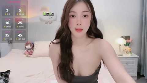 Media: Video of an East Asian woman with long brown hair, light skin, and a slender build, wearing a black strapless top, sitting on a bed with white sheets. The room features a digital clock and stuffed toys in the background.