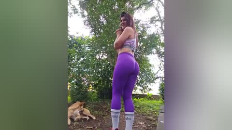 Media: Video of a curvy woman with medium skin tone, wearing tight purple leggings and a sports bra, standing in a wooded area with a golden retriever and lush greenery.