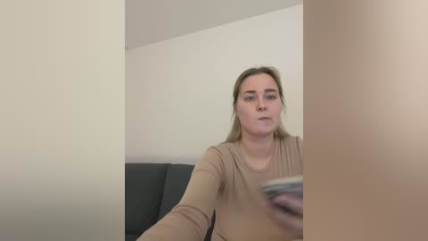 Media: Video of a Caucasian woman with light skin and brown hair, wearing a beige long-sleeve top, sitting on a dark gray couch against a plain white wall. She is holding a smartphone in her right hand.