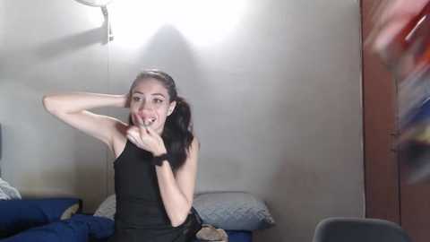 Media: Video of a slender, dark-haired woman in a black tank top, sitting on a bed with blue sheets, making a finger gun gesture in a dimly lit room.