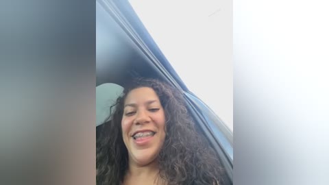 Media: Video of a smiling woman with curly dark hair, braces, and medium skin tone, sitting in a car. The background is a blurry white wall.