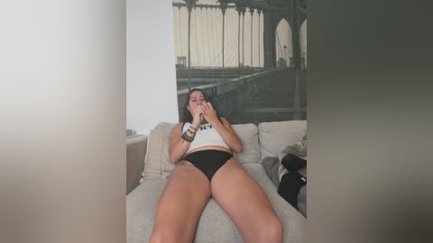 Media: Video of a young woman with long brown hair, wearing a white crop top and black panties, lying on a light-colored couch, looking at her phone. Background features a monochrome wall mural of a bridge.