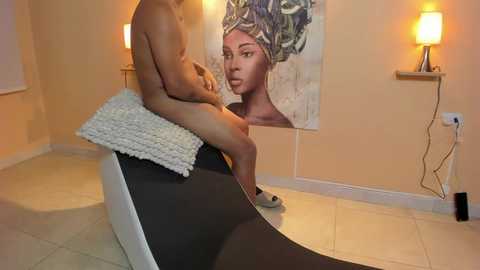 Media: Video of a nude, muscular Black man sitting on a black couch, wearing a white knitted blanket, with a large portrait of a Black woman with braided hair on beige walls, warm lighting, and modern decor.