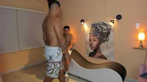 Media: Video of a muscular shirtless man wearing white shorts with a Louis Vuitton logo, carrying a woman with short hair in a lounge room with a chaise, large portrait, and warm lighting.