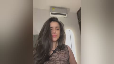 Media: Video of a young woman with long black hair, wearing a sheer black lace top, standing in a dimly lit room with beige walls, an air conditioning unit on the ceiling, and a blurred background.