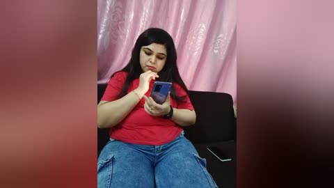 Media: Video of a plus-sized South Asian woman with long black hair, wearing a red top and blue jeans, sitting on a black couch, absorbed in her smartphone, against pink floral curtains.