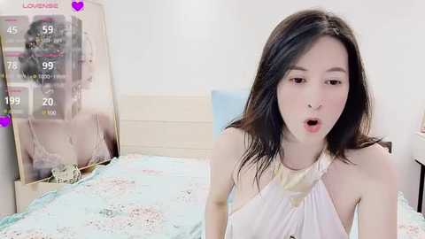 Media: A video of a young Asian woman with long black hair, wearing a white halter top, lying on a bed with floral sheets, with a \"Lovense\" screen in the background.